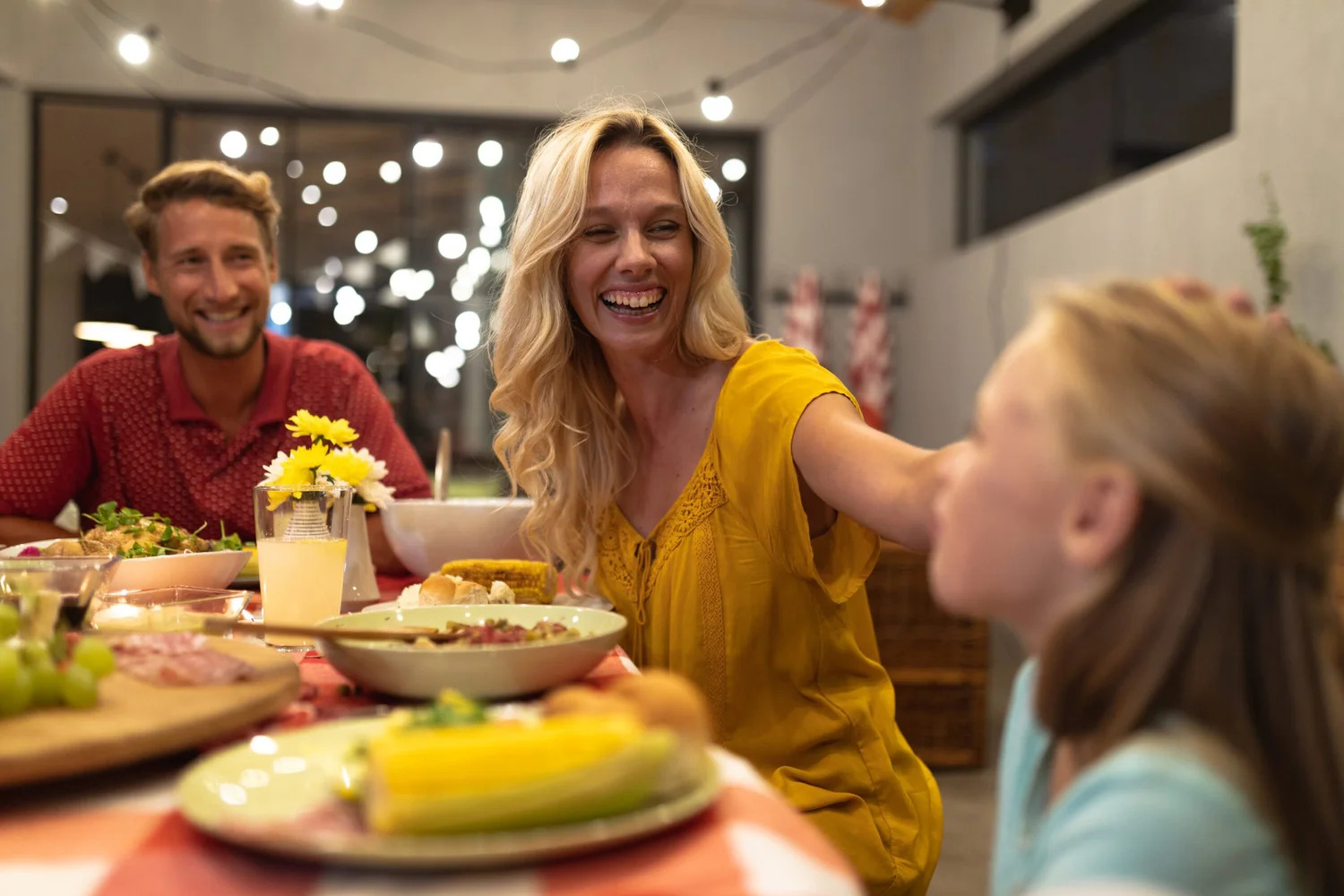 A family laughing together around a dinner table of salads, corn and shared dishes under string lights